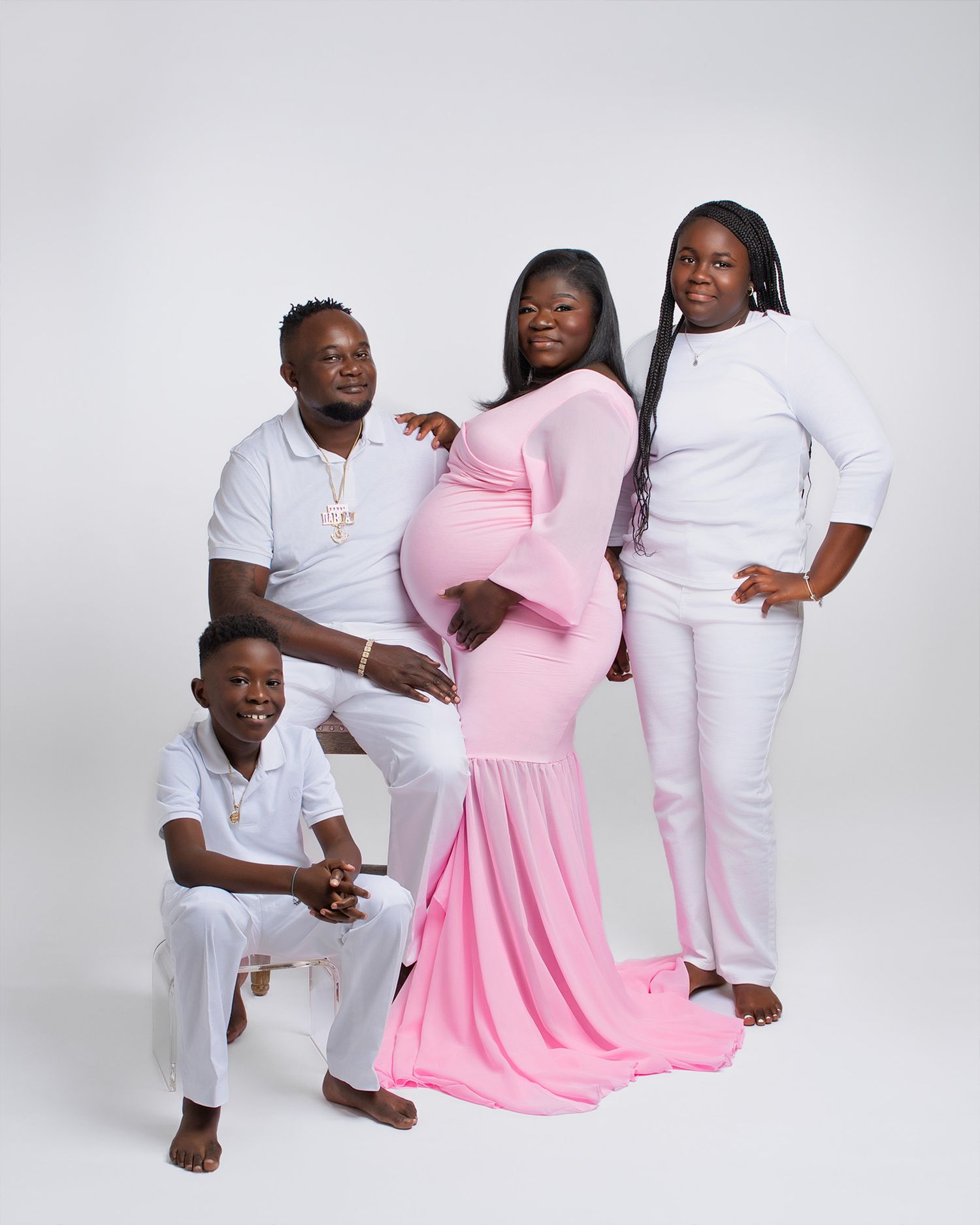 Family maternity photo with group wearing coordinated white outfits and pink gown against light background.