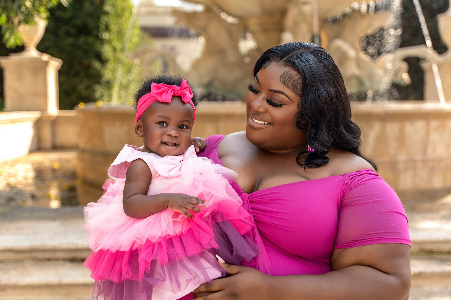 Adult and baby coordinate in pink outfits while posing by outdoor fountain.