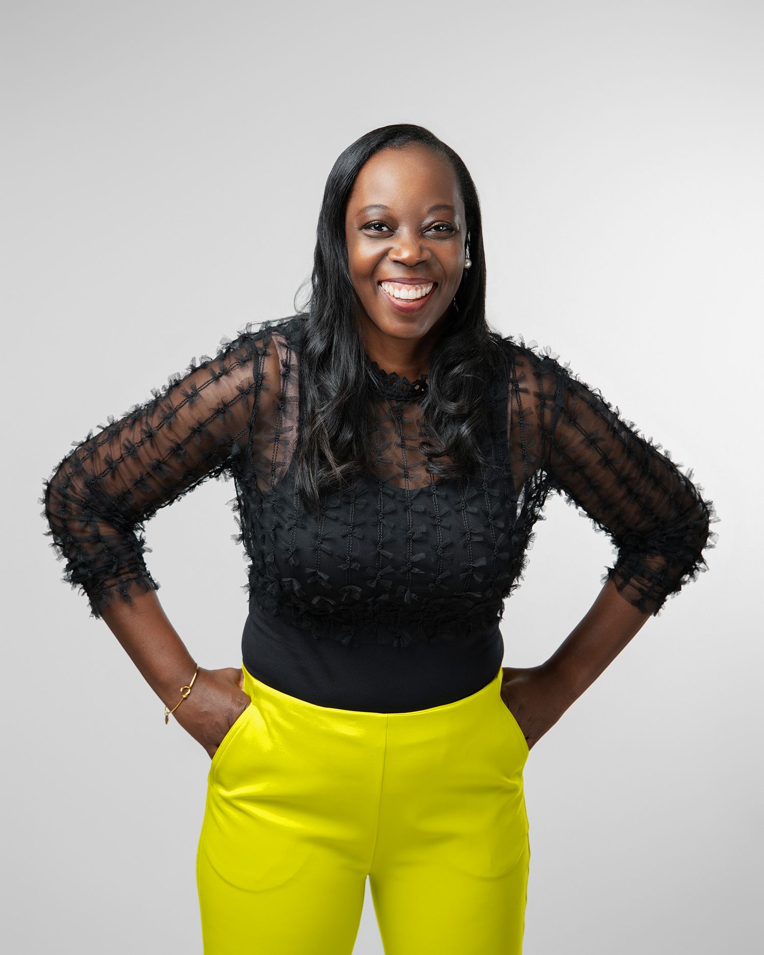 Studio portrait of someone in black lace top and bright yellow pants against light background.