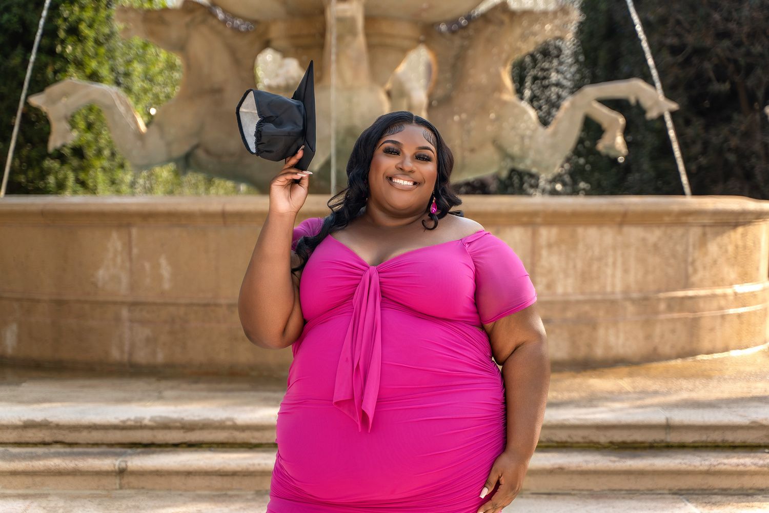 Woman in bright pink dress poses confidently in front of an ornate outdoor fountain.