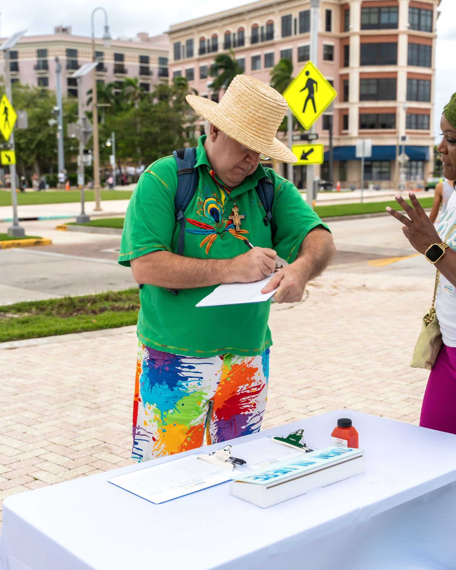 Person in green shirt and colorful shorts taking notes at outdoor event registration table.
