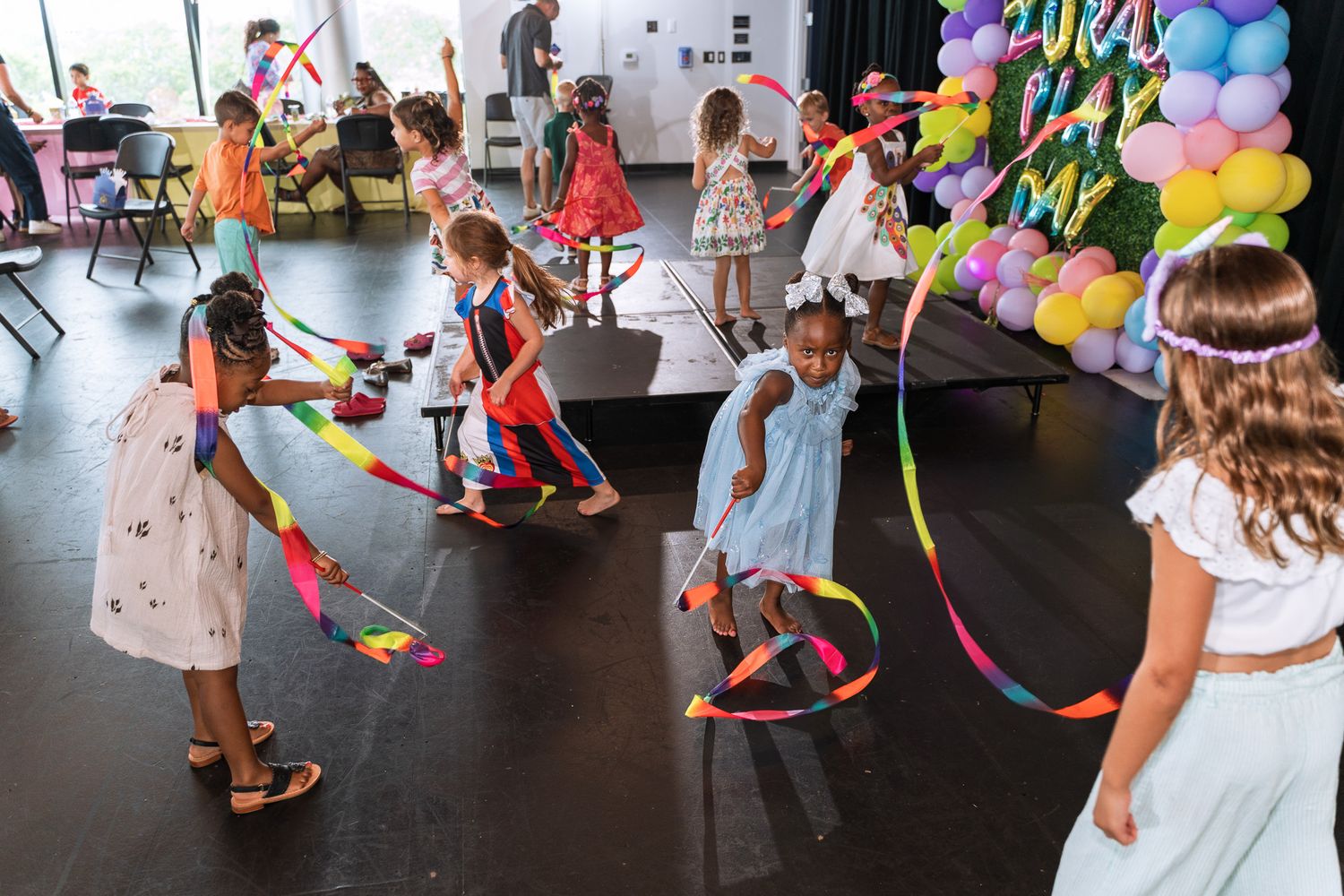 Children playing with colorful ribbons and hula hoops at a party with balloon decorations.