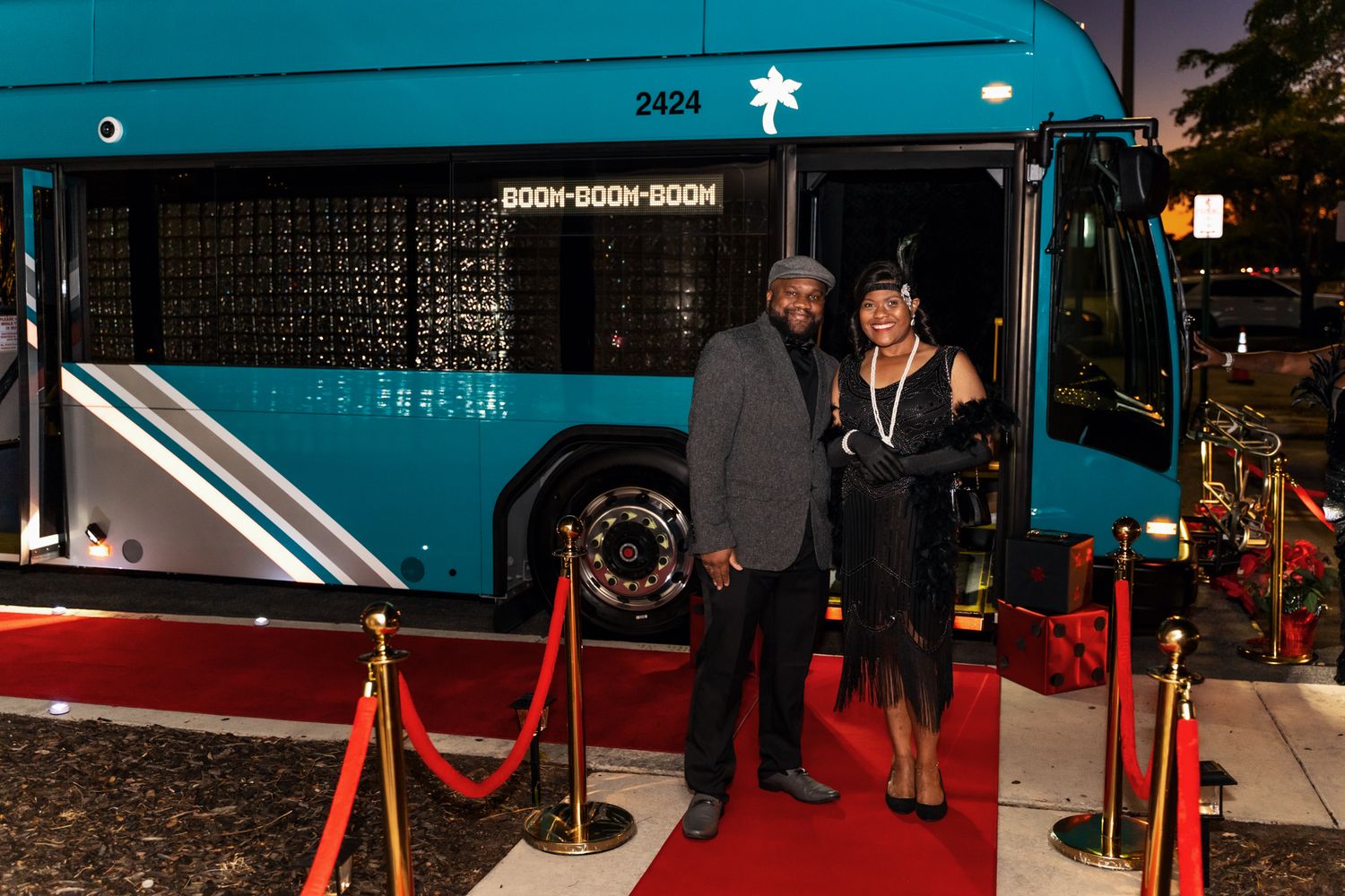 An elegant couple poses on a red carpet in front of a turquoise bus at night.