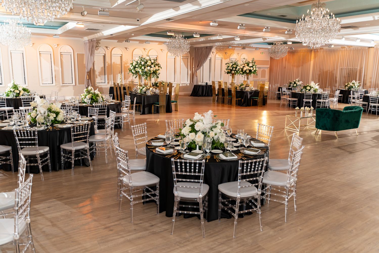An elegant ballroom with crystal chandeliers, round tables with black linens, and silver chiavari chairs arranged for a formal event.