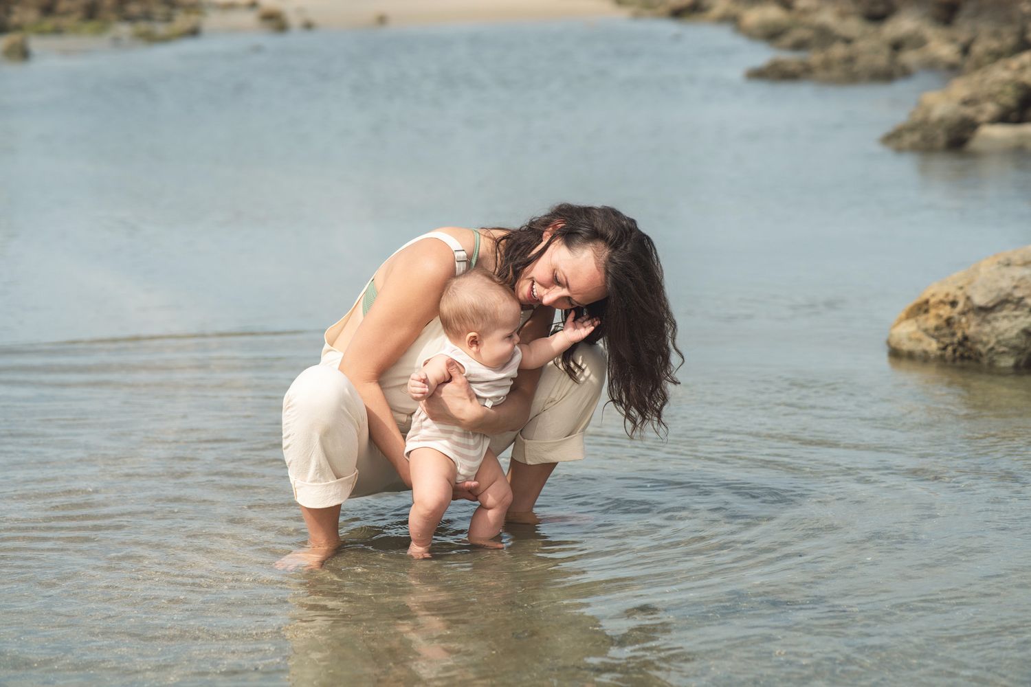 A parent and baby play together in shallow coastal waters surrounded by rocky outcrops on a sunny day.