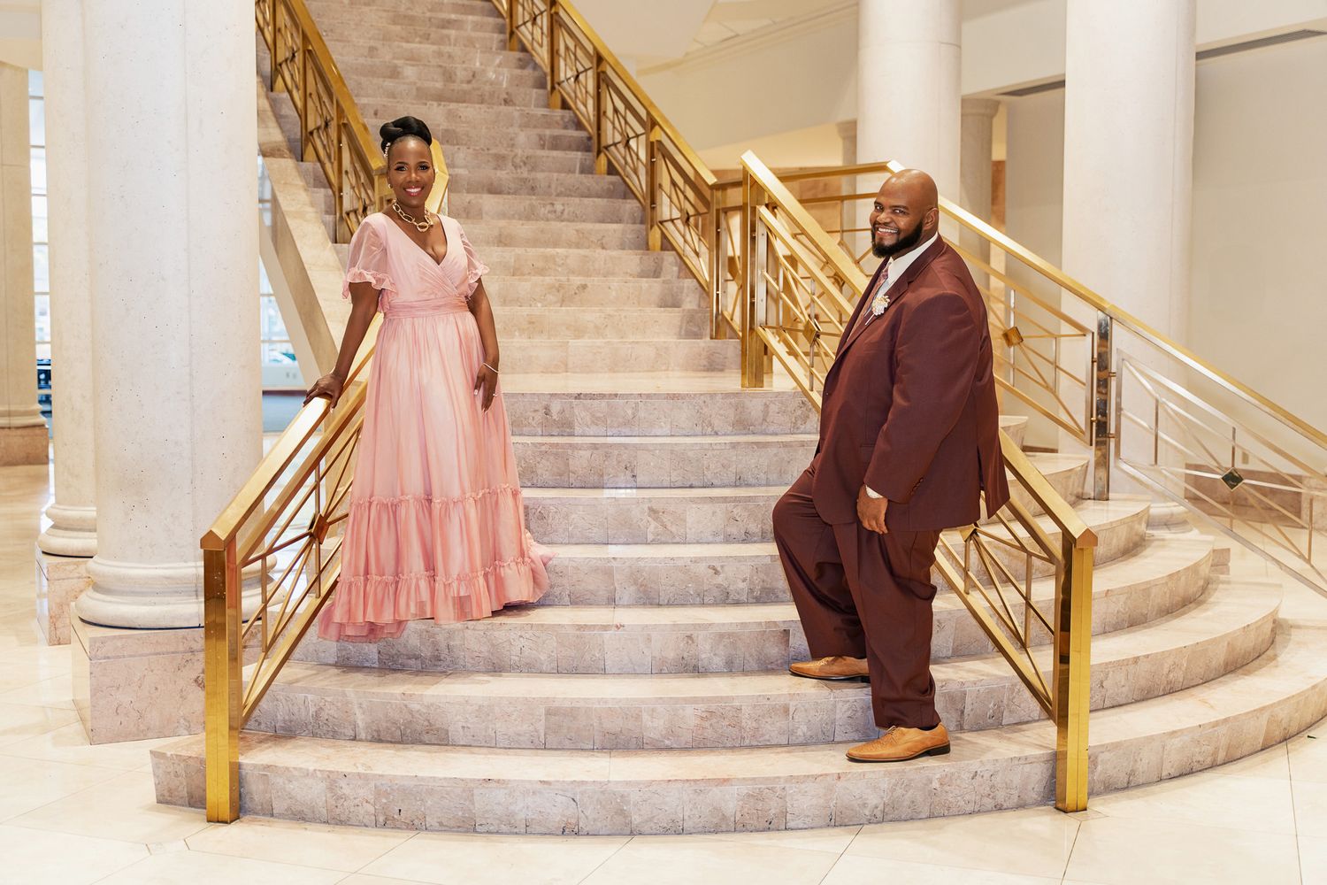 A couple stands apart on elegant marble stairs with gold railings in formal wedding attire.