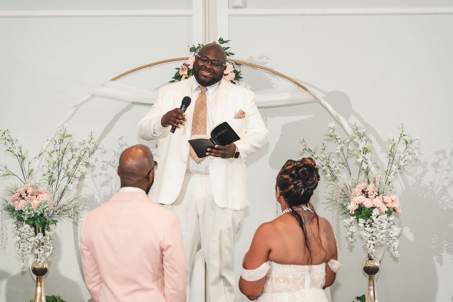 Wedding ceremony in progress with an officiant in white and decorative floral arch.