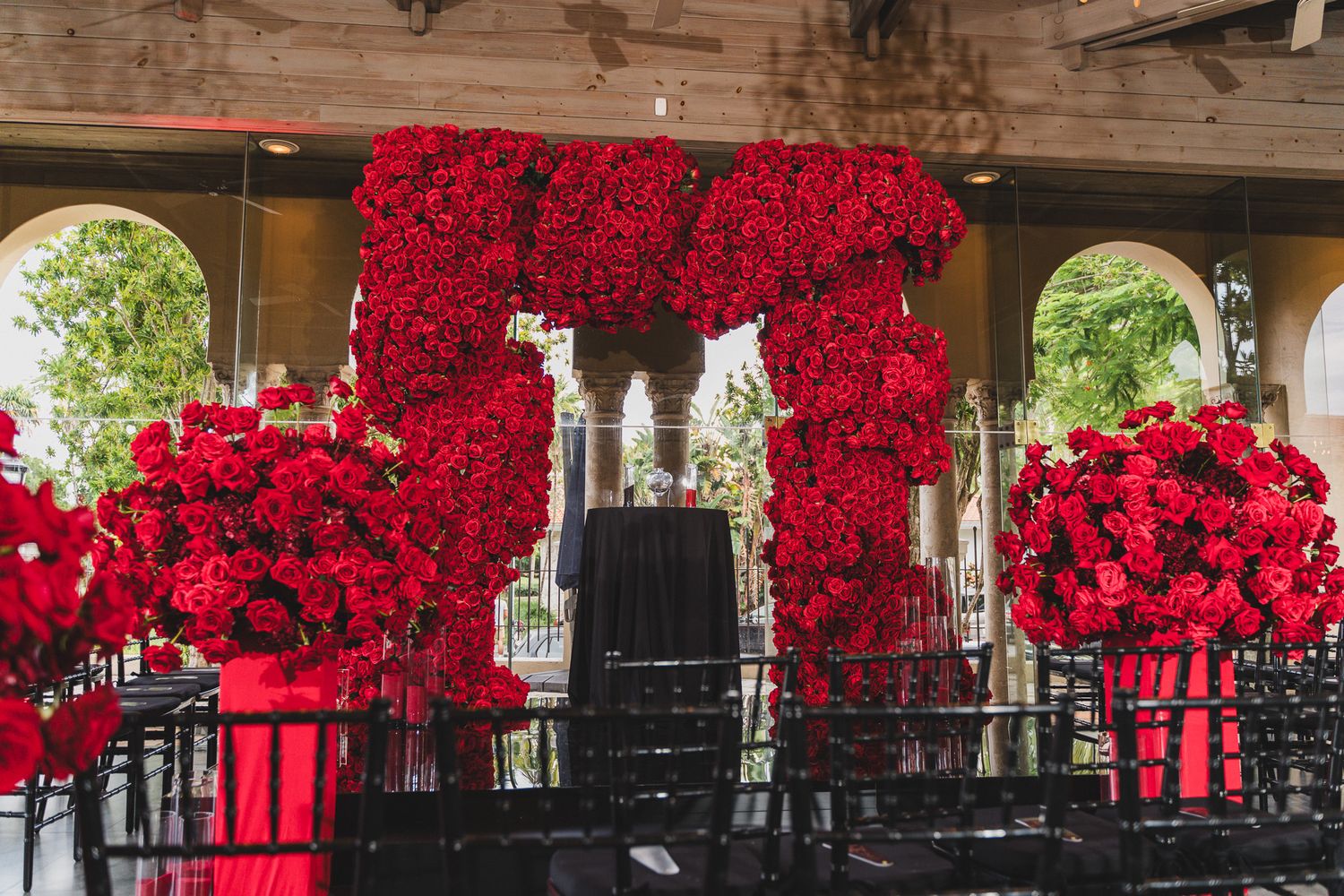Luxurious wedding ceremony setup with dramatic red rose floral arch and black chairs.