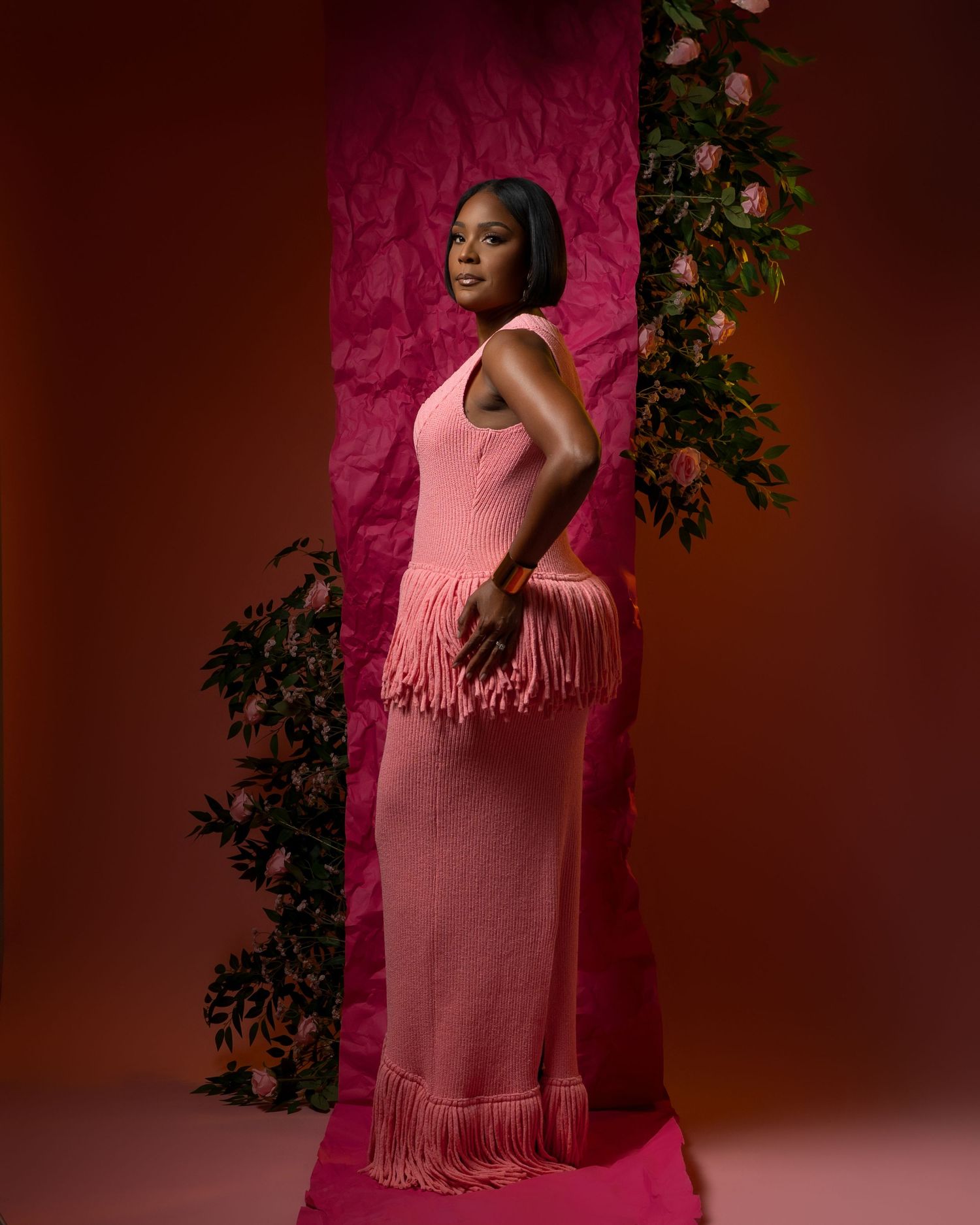 A figure in a pink fringe dress poses against a pink backdrop with green foliage accents in a fashion photoshoot.
