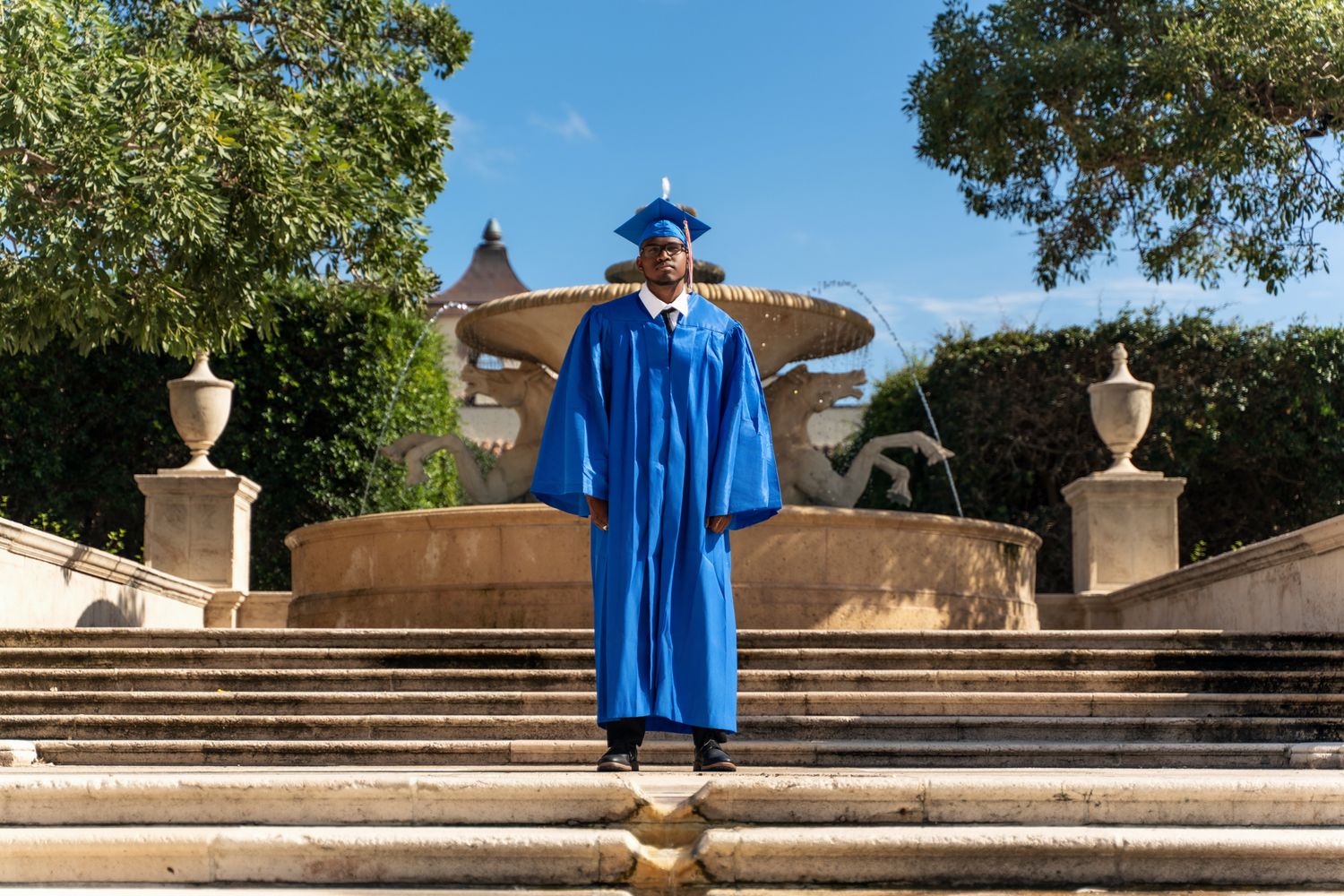 Graduate in bright blue cap and gown standing on grand stone steps with decorative urns in a sunny outdoor setting.