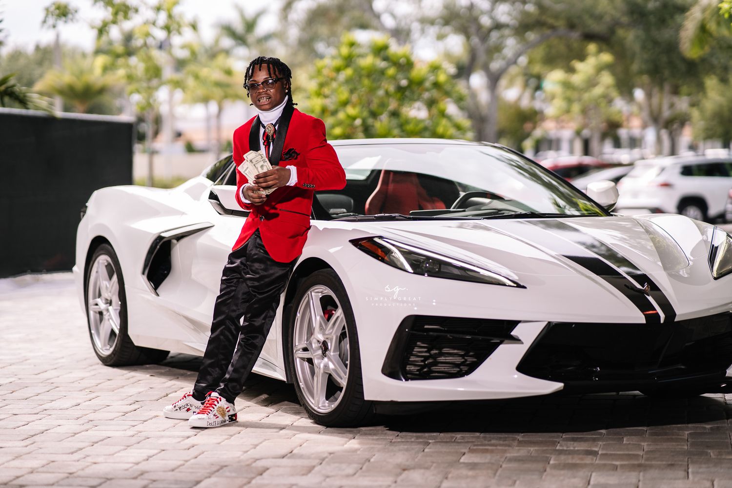 A person in red attire poses beside a white Corvette C8 sports car.