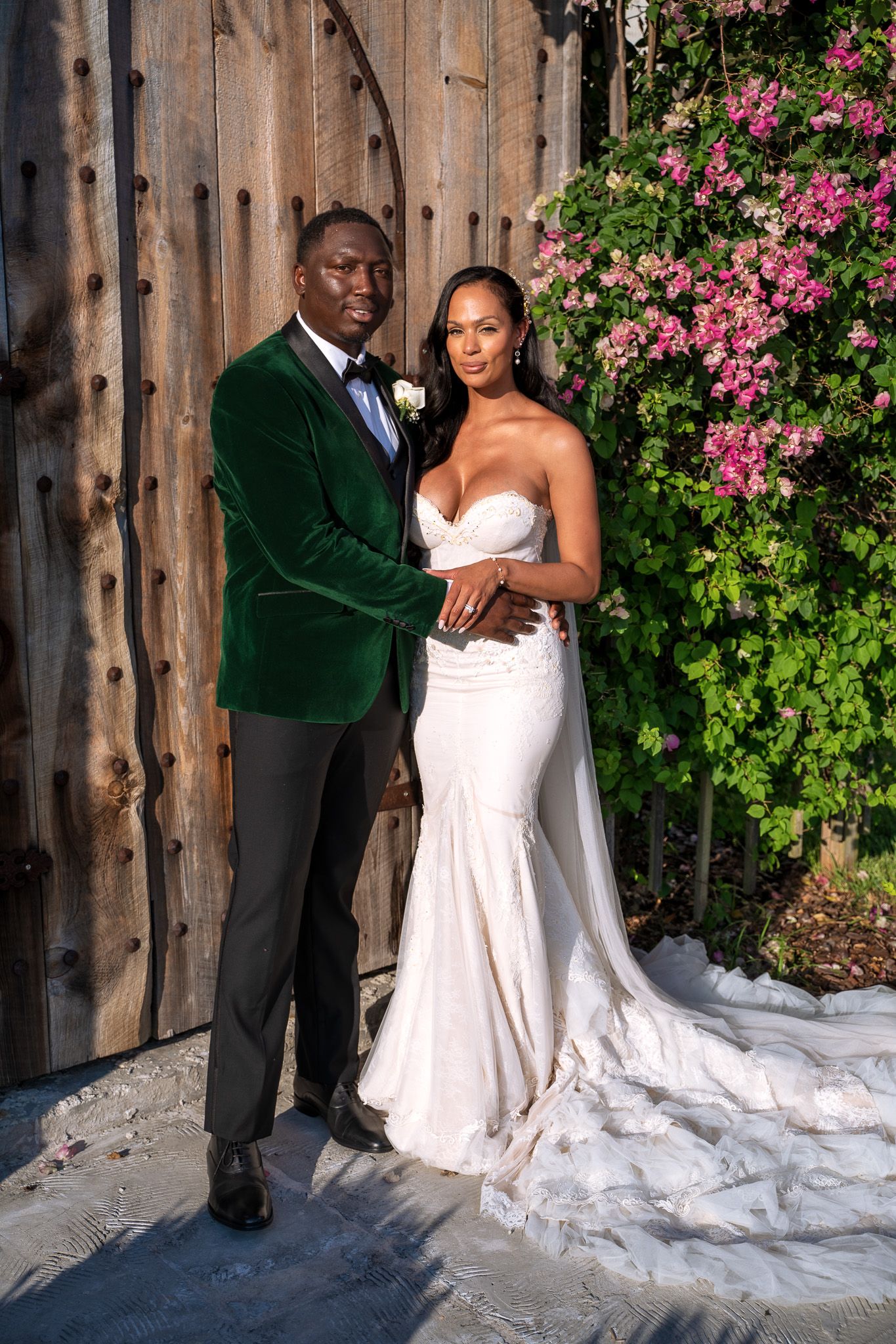 Wedding couple poses against rustic wooden wall with pink flowering vine for romantic portrait.