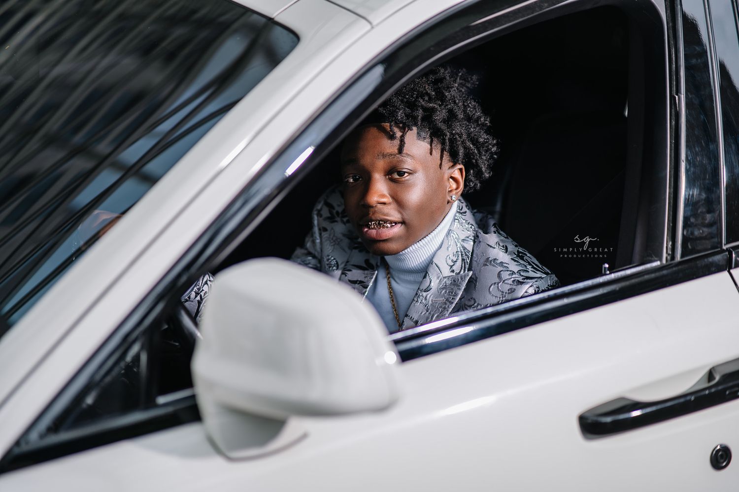 Person wearing gray patterned suit smiling while sitting in white car.
