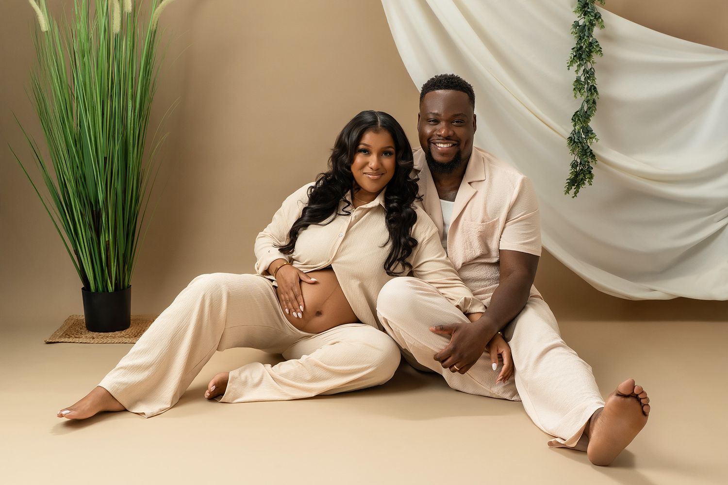 Expectant couple in coordinating cream outfits pose on floor during maternity photoshoot.