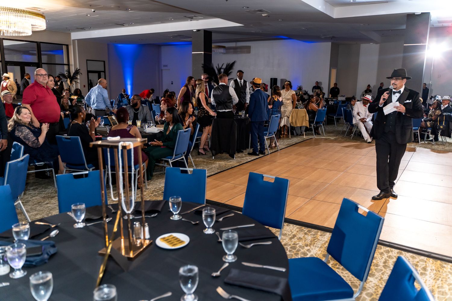 An energetic crowd gathers in a ballroom with blue chairs and formal table settings.