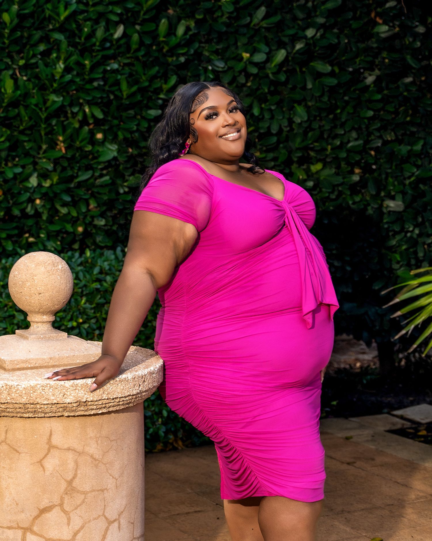 Woman wearing form-fitting hot pink dress poses elegantly against garden foliage.