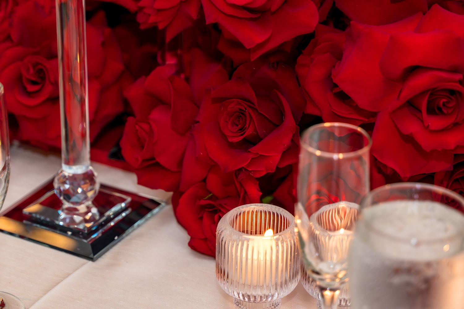 Red roses and glowing candles create an elegant romantic table setting with crystal glassware against a dark background.