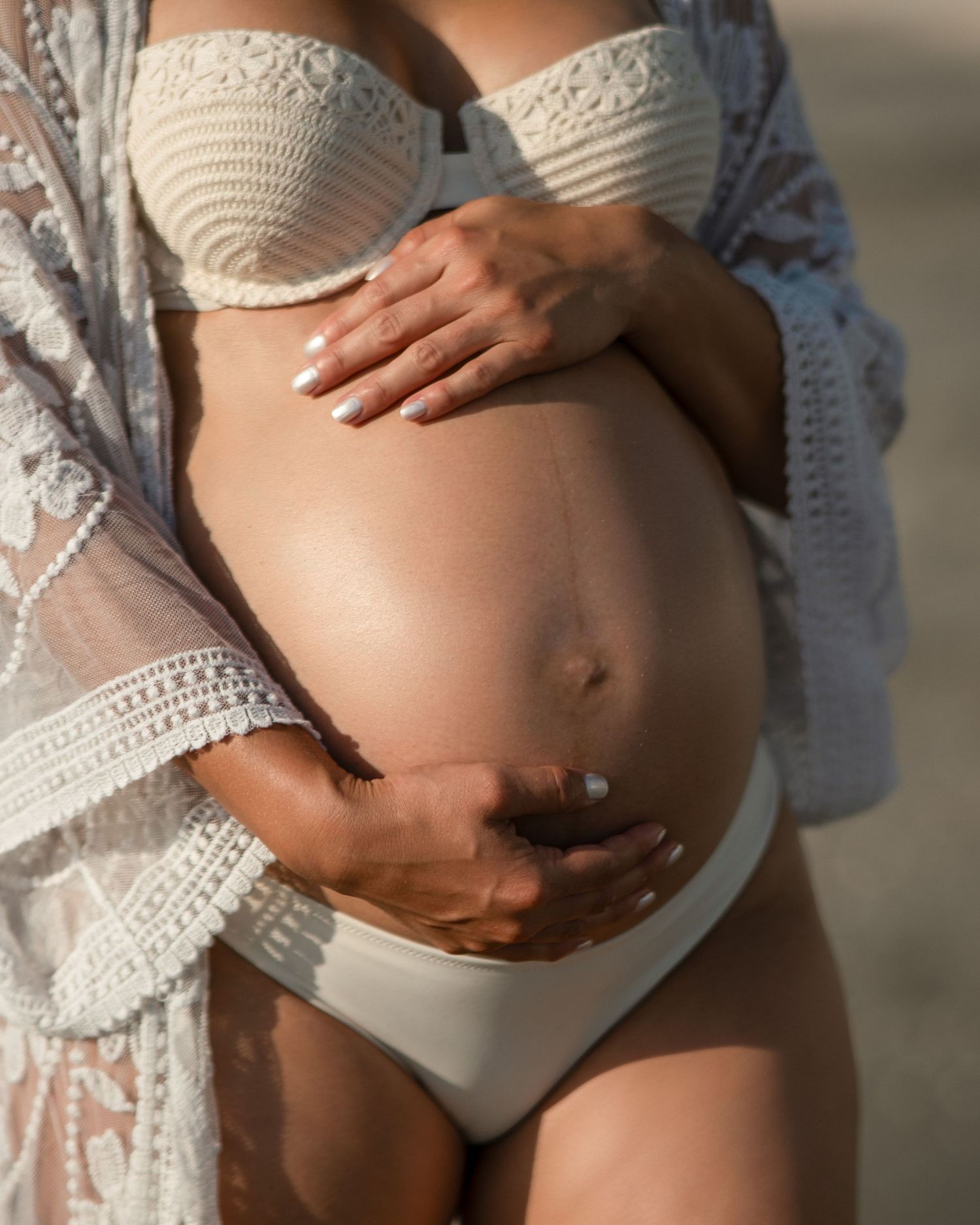 Close-up of pregnant belly with hands gently cradling bump, wearing white lace and swimwear.