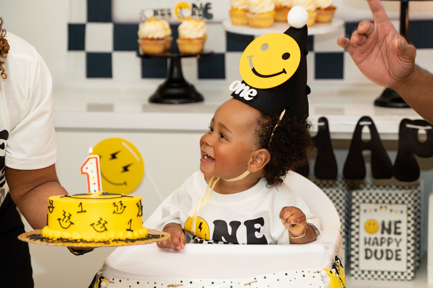 A child celebrates their first birthday with a smiley face themed cake and party hat.