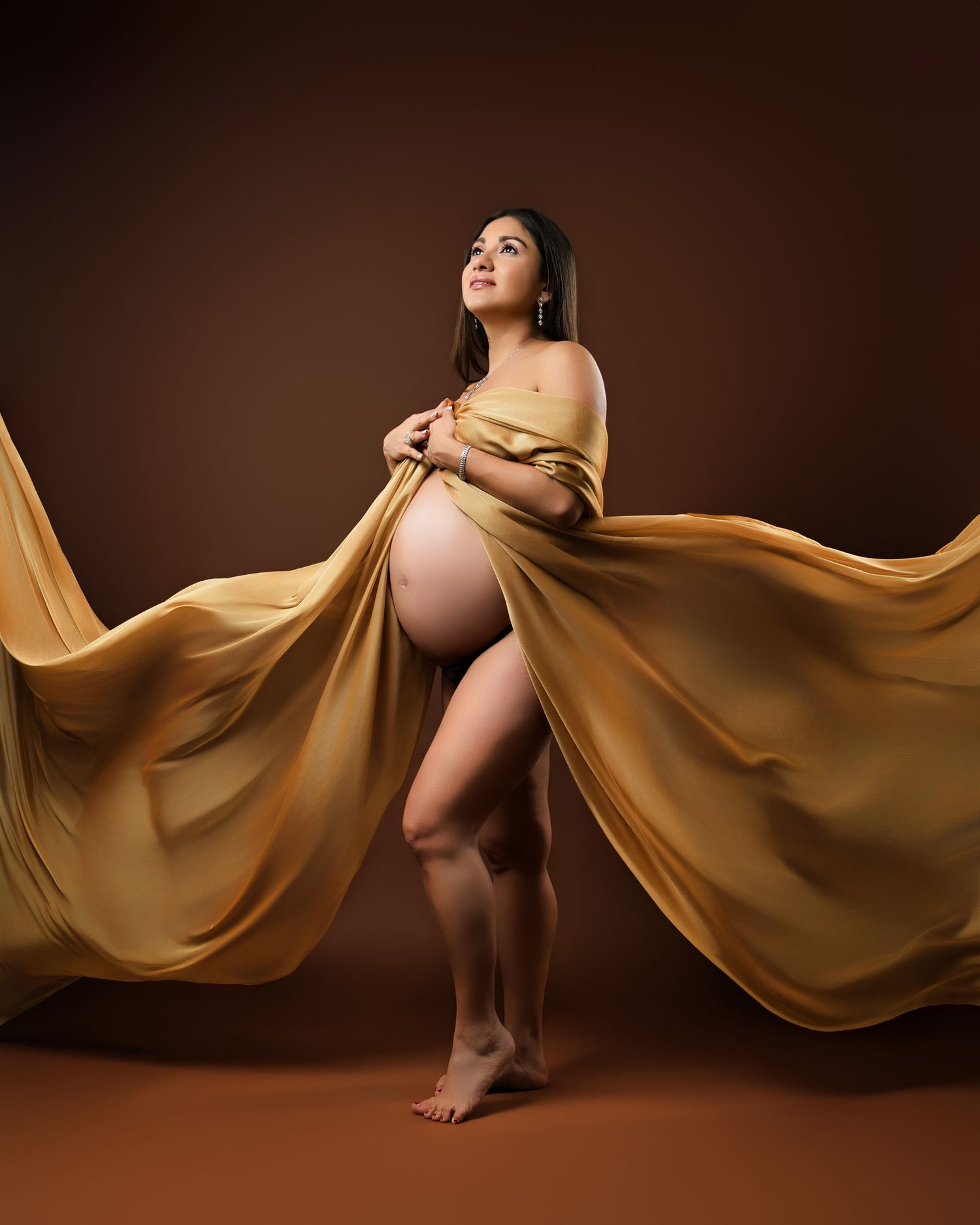 Dynamic maternity photo with flowing gold fabric creating dramatic movement against brown backdrop.
