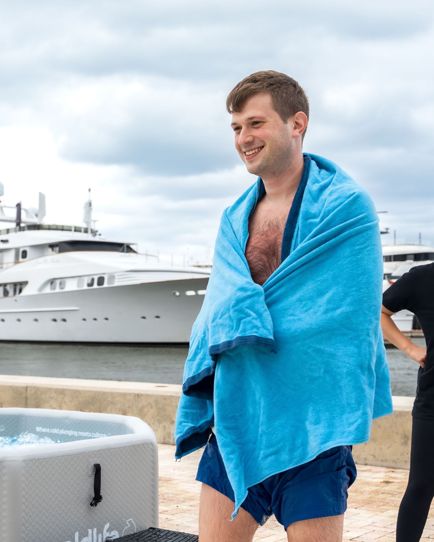 Someone wrapped in a blue towel stands near a luxury yacht at a marina.