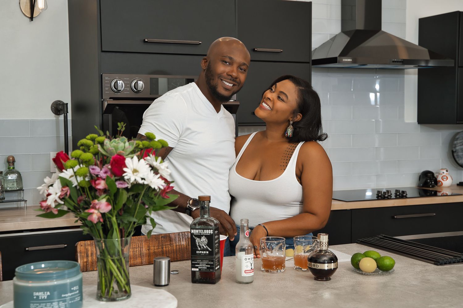 A couple shares an intimate moment in a modern kitchen with flowers and beverages on the counter.