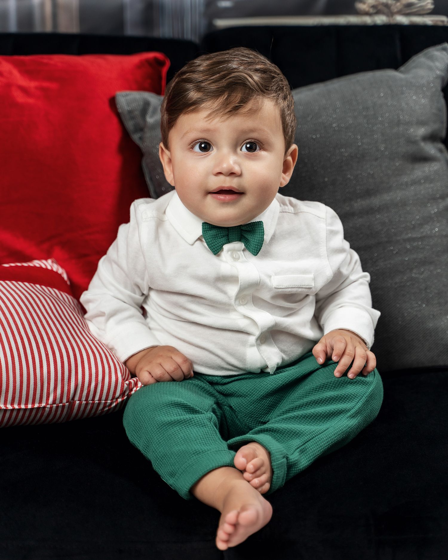 Baby in formal white shirt and green bow tie sitting on couch with red and grey pillows.