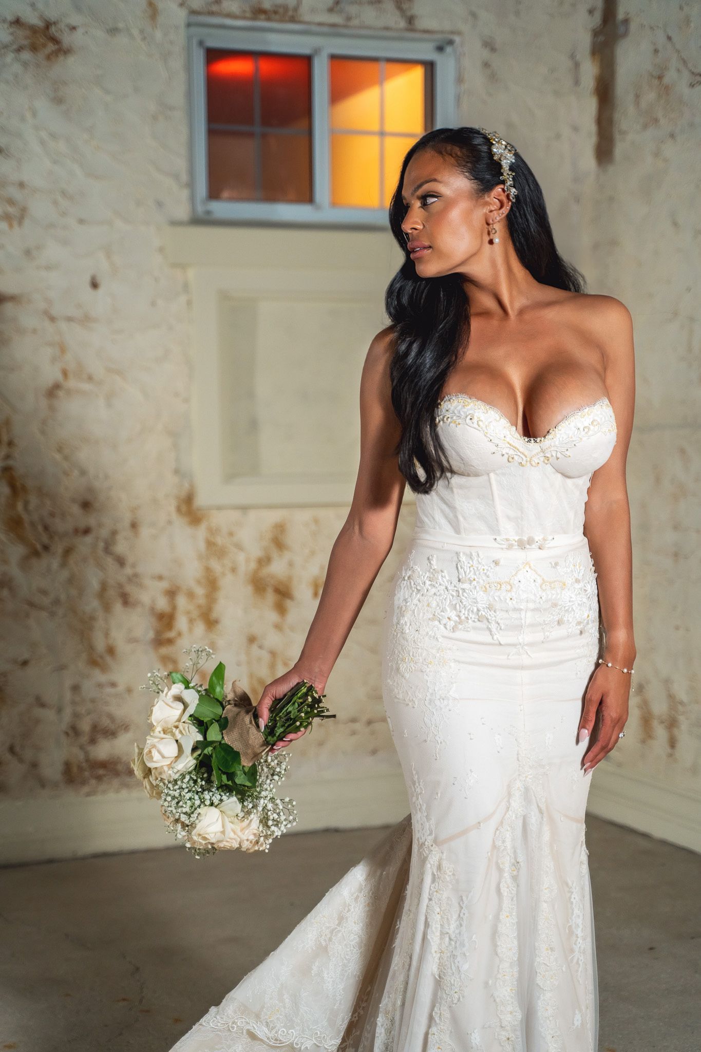 Bride poses in fitted lace wedding dress holding white floral bouquet against textured wall.