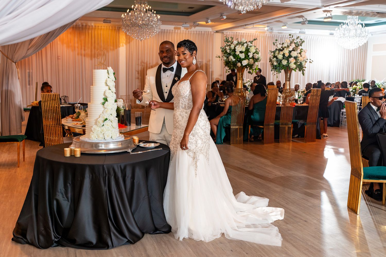 A romantic wedding scene with a couple posing next to their elegant tiered white cake at an indoor reception venue.
