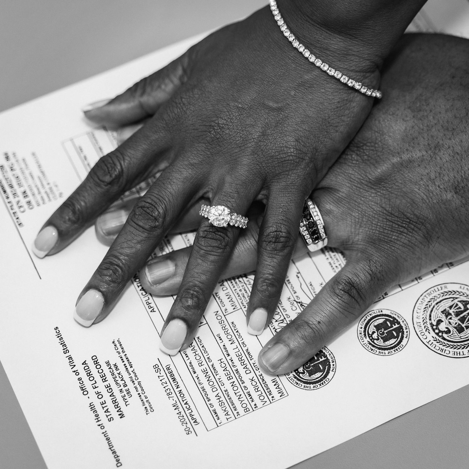 Black and white close-up of hands with wedding rings on official documents.