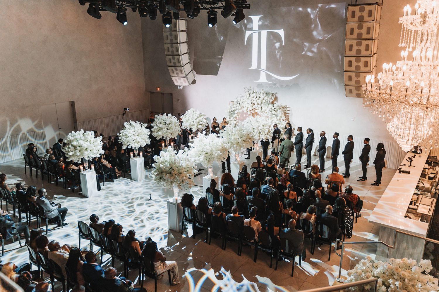 Aerial view of an elegant indoor wedding ceremony with white floral decorations and gathered guests.