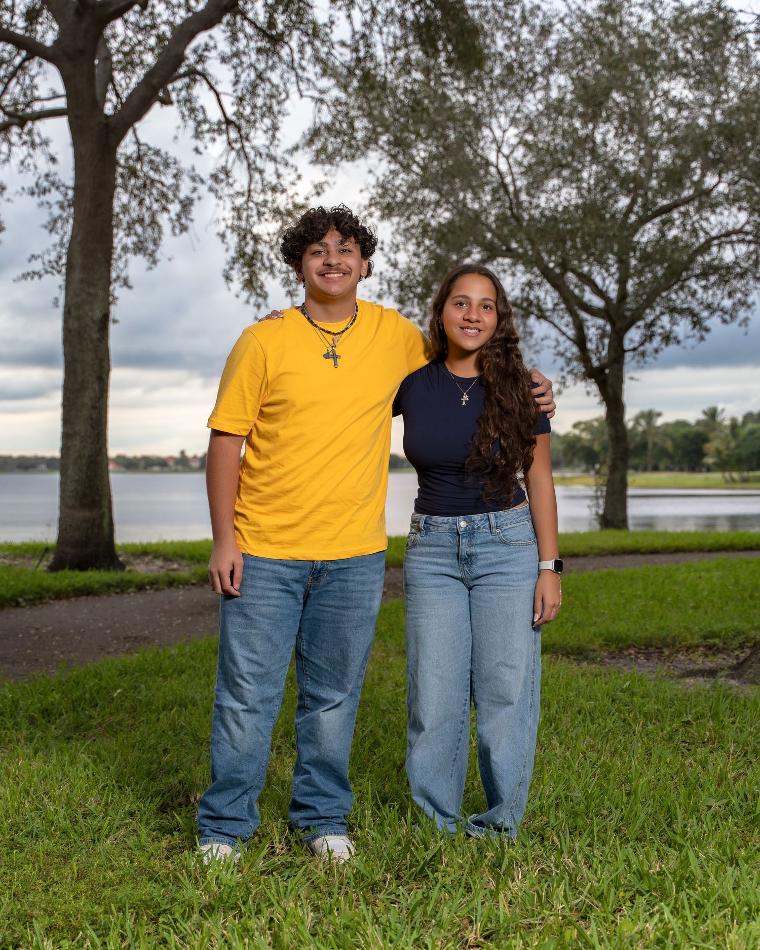 Two people in yellow and navy outfits stand together on green grass near a waterfront with trees in the background.