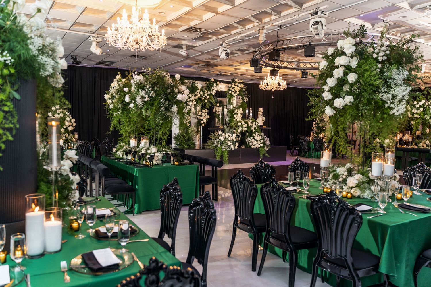 Elegant indoor wedding reception venue with emerald green tablecloths, crystal chandeliers, and lush floral arrangements.