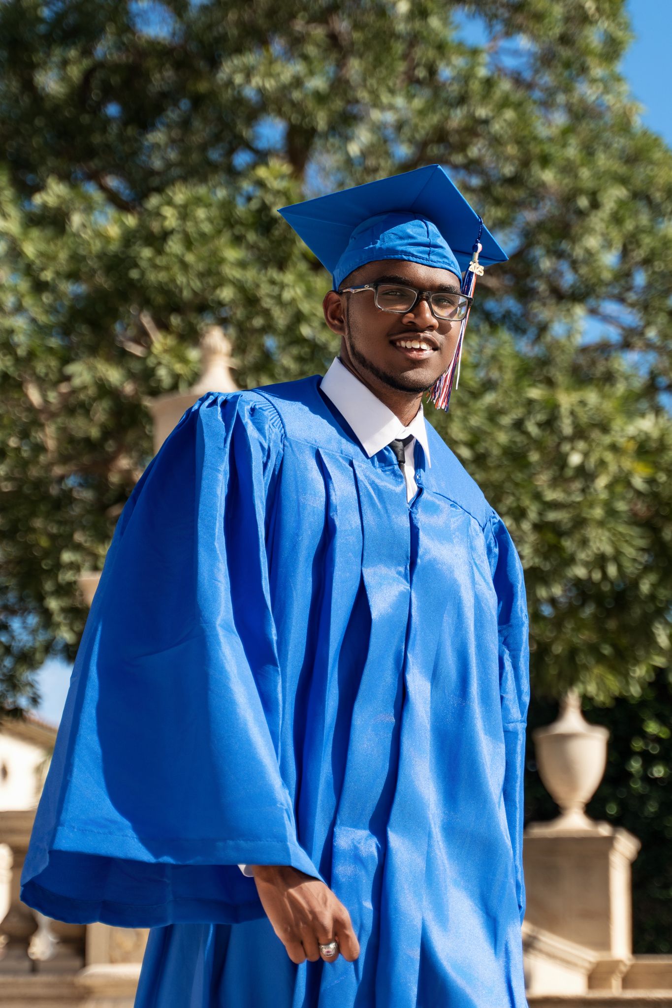 Graduate in bright blue cap and gown stands proudly outdoors during commencement celebration.