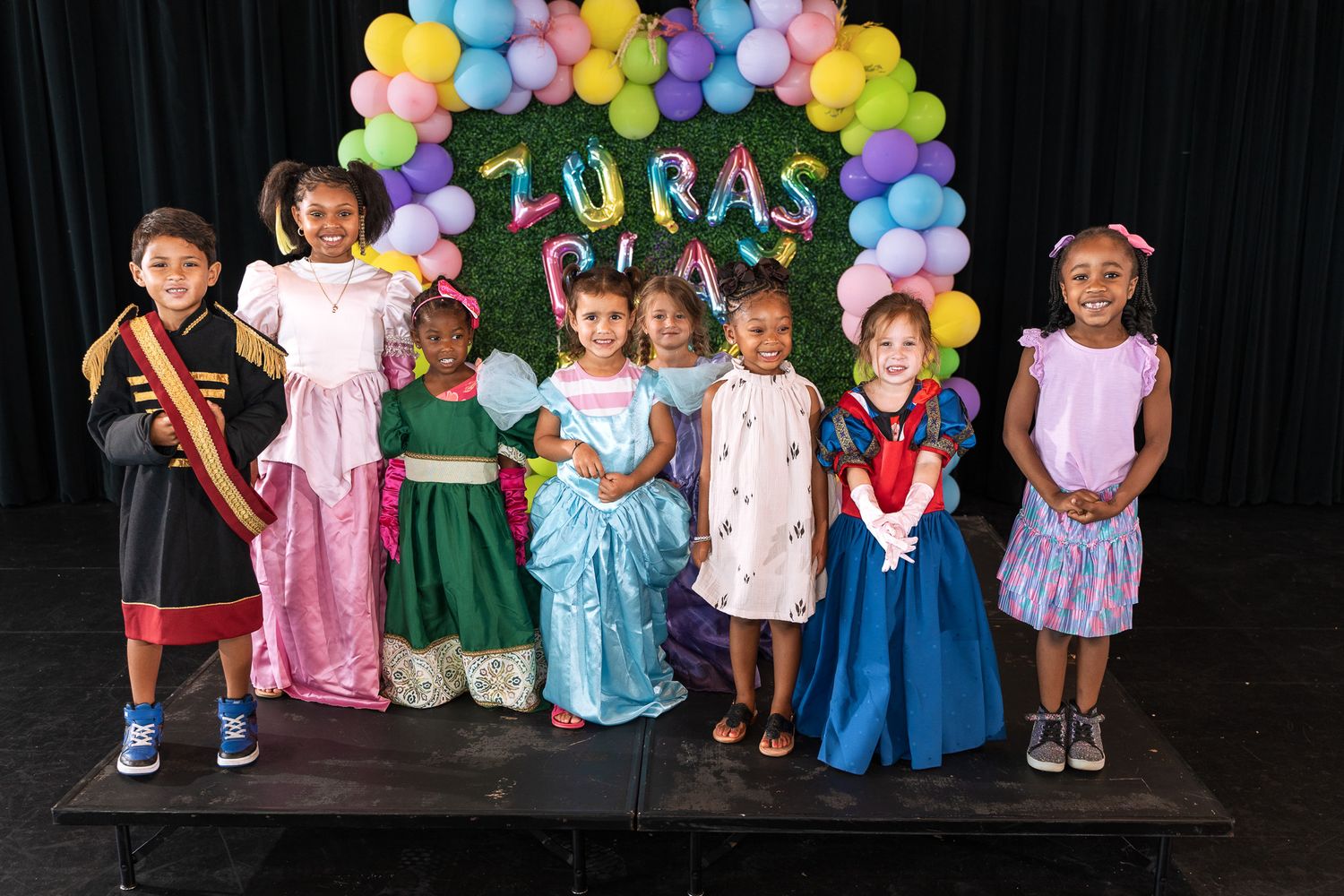 Children in costumes pose under colorful balloon arch at party celebration.