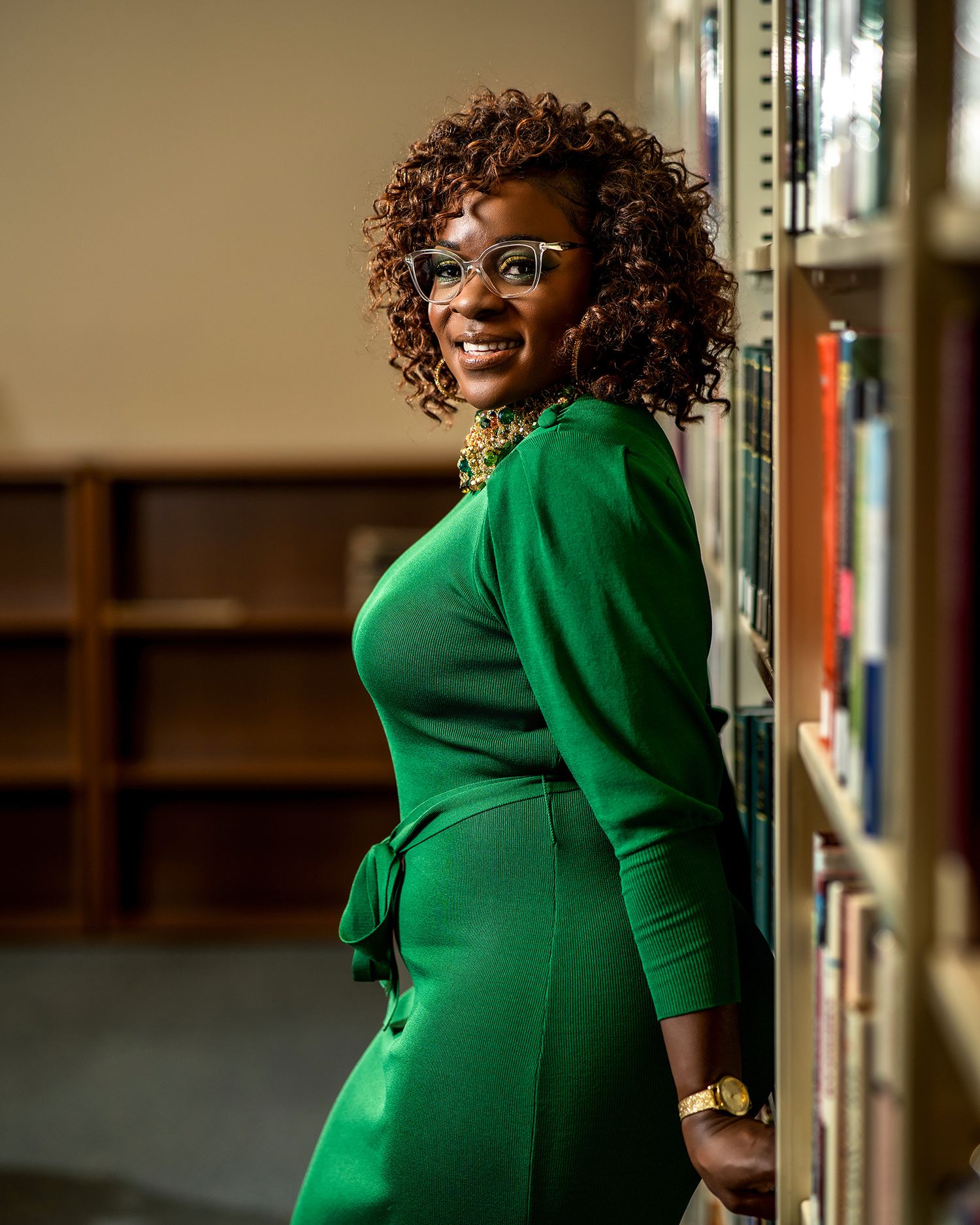 Graduate in stylish green dress and glasses poses by library shelves.