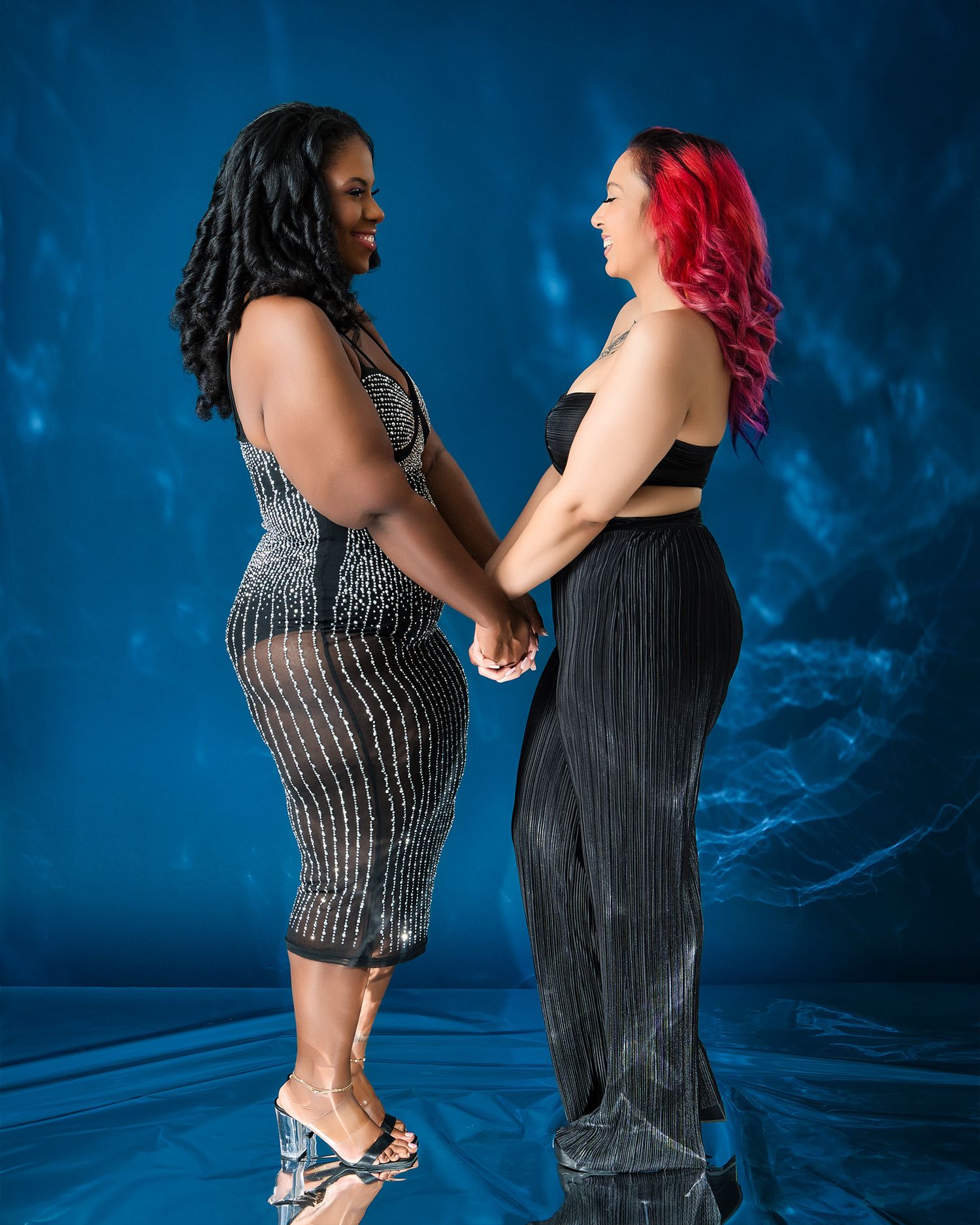 Two people in elegant dresses holding hands against blue studio backdrop.