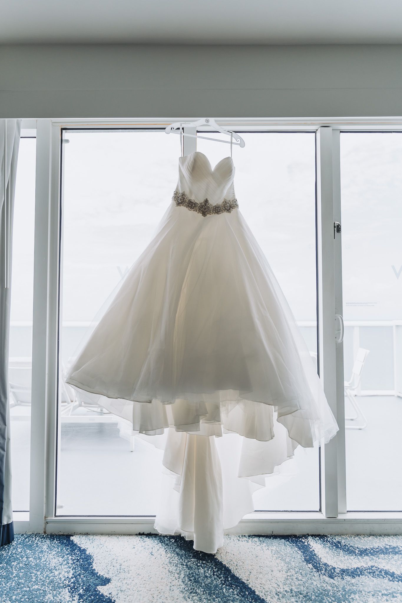 White wedding gown with crystal belt detail hanging in front of bright windows.