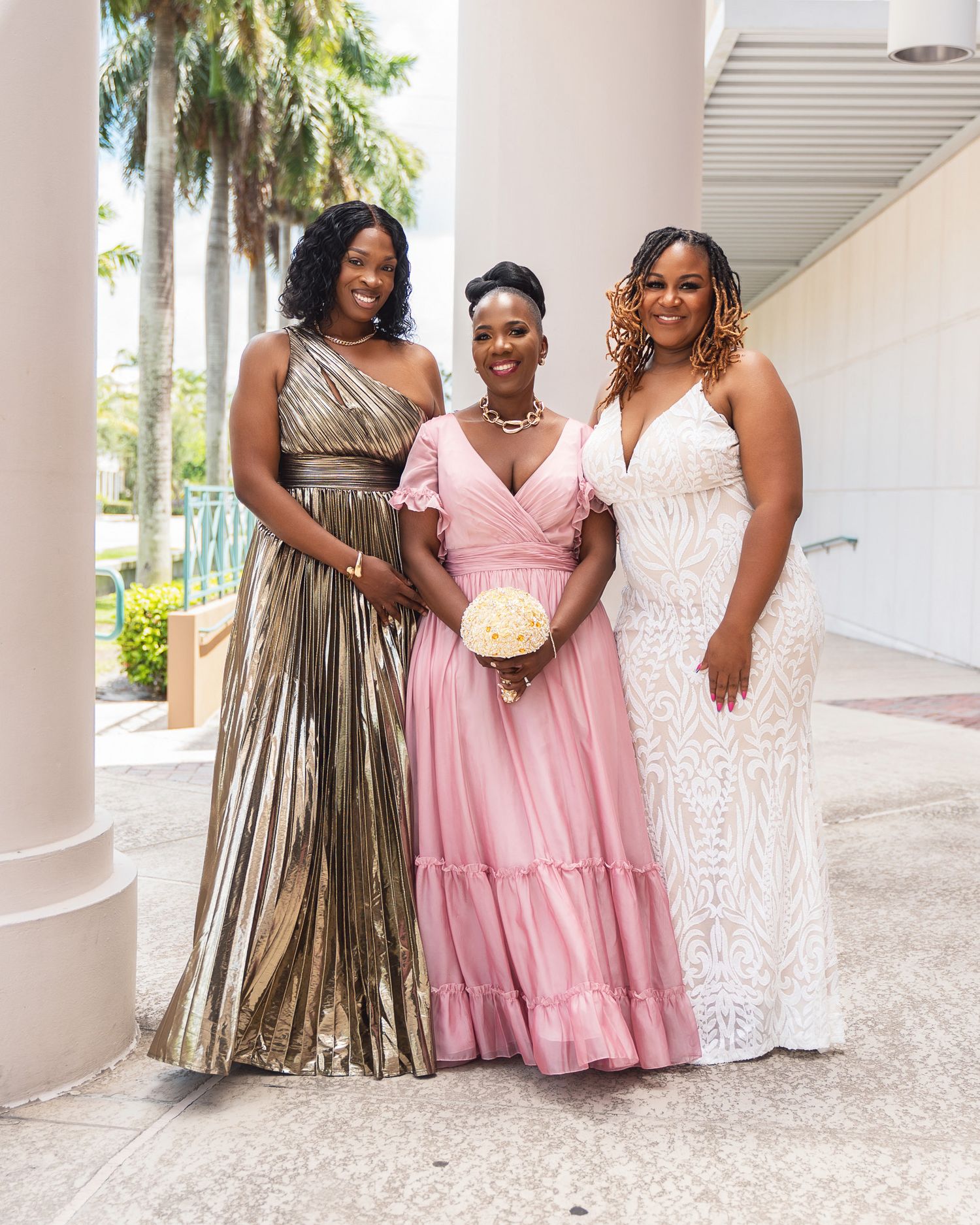 Three women in formal dresses pose together against white columns and palm trees.