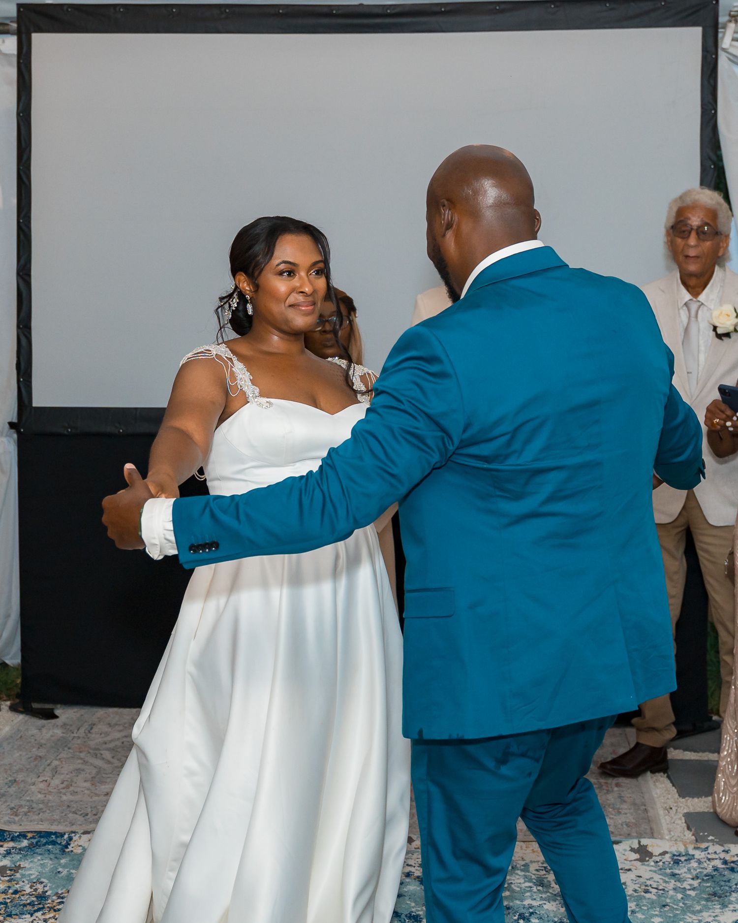 Couple enjoys their first dance at wedding reception with guests and backdrop behind them.