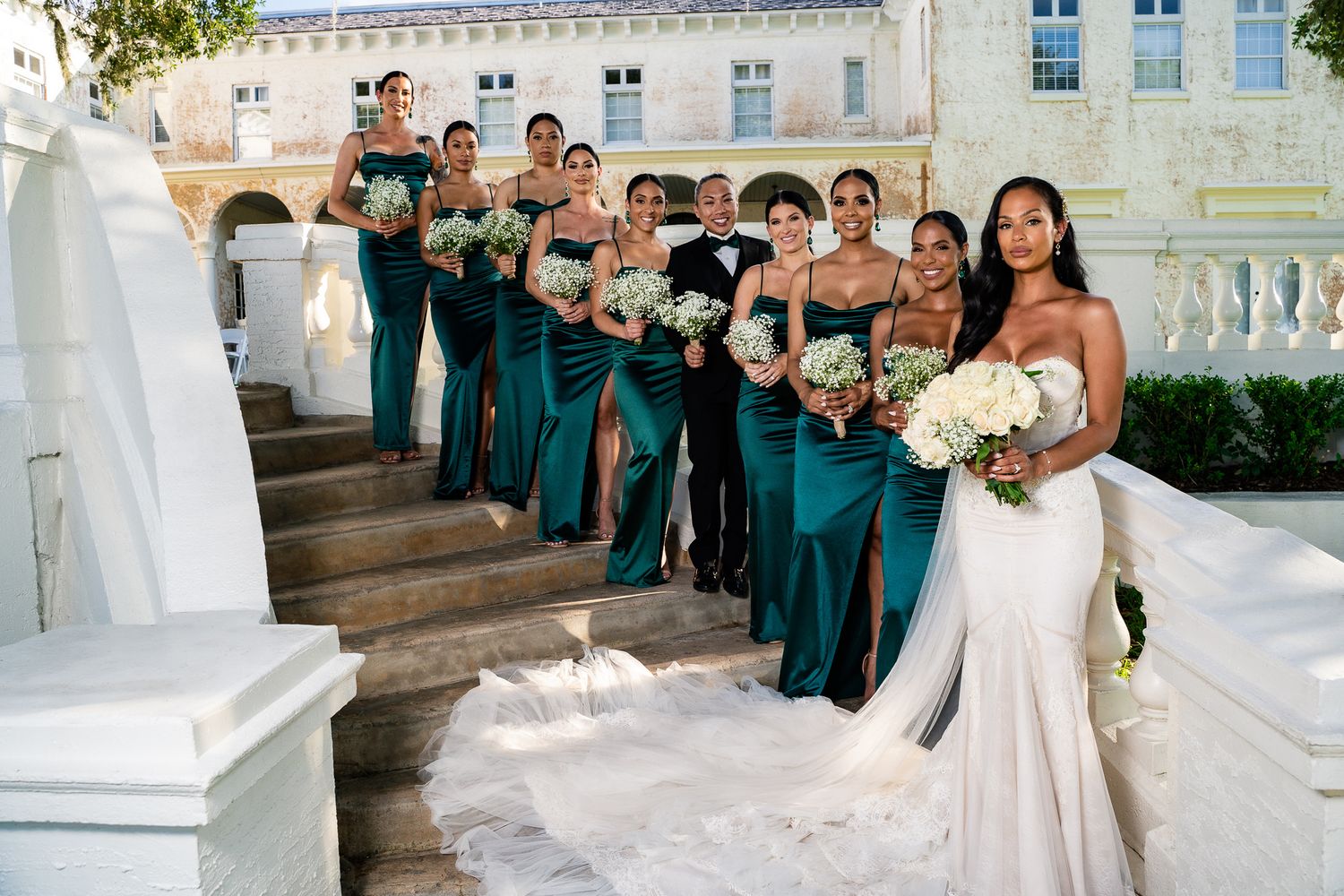 Bridesmaids in teal dresses pose with bride in white gown on elegant stone steps.
