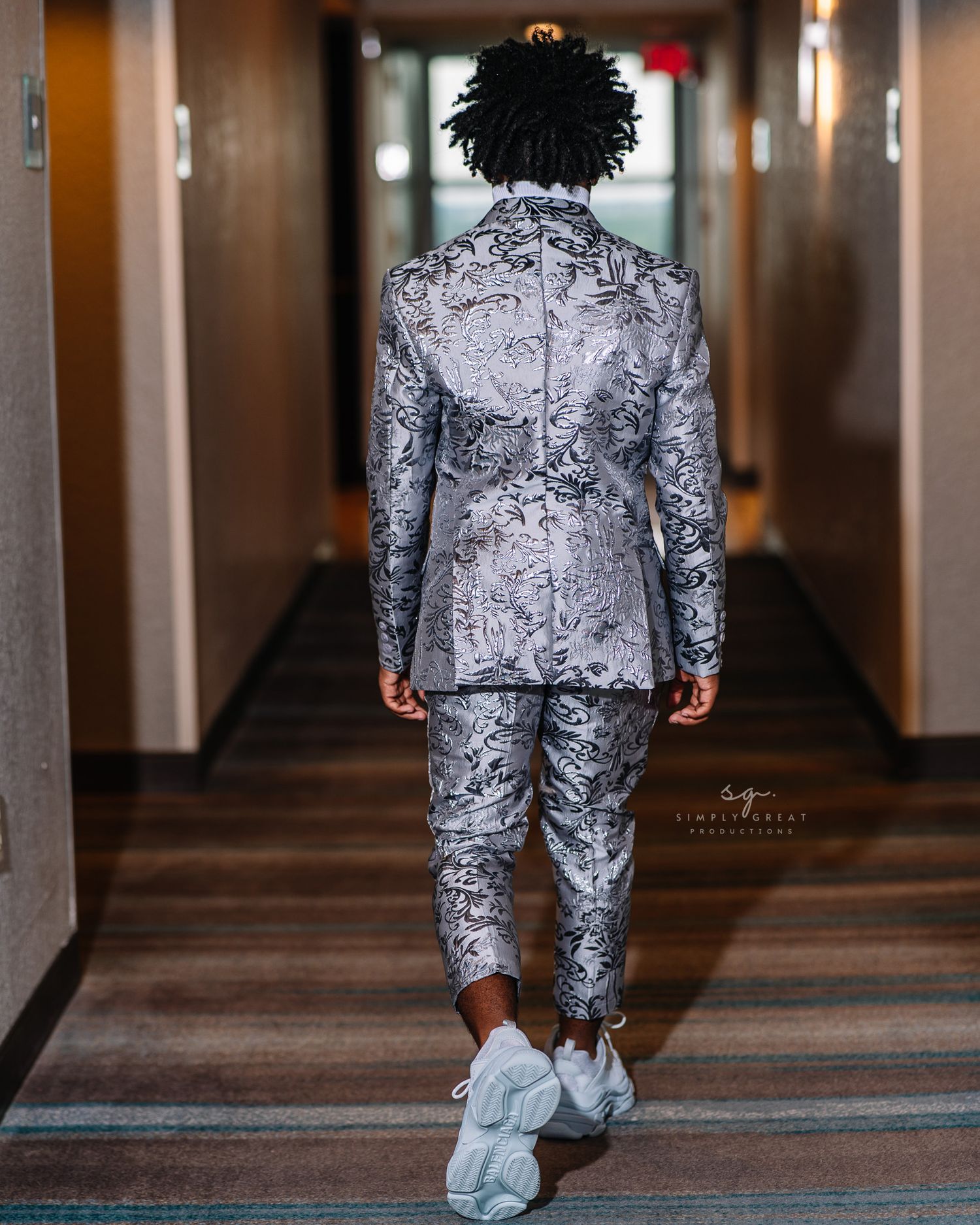 Back view of someone wearing a silver patterned suit and white sneakers in a hallway.