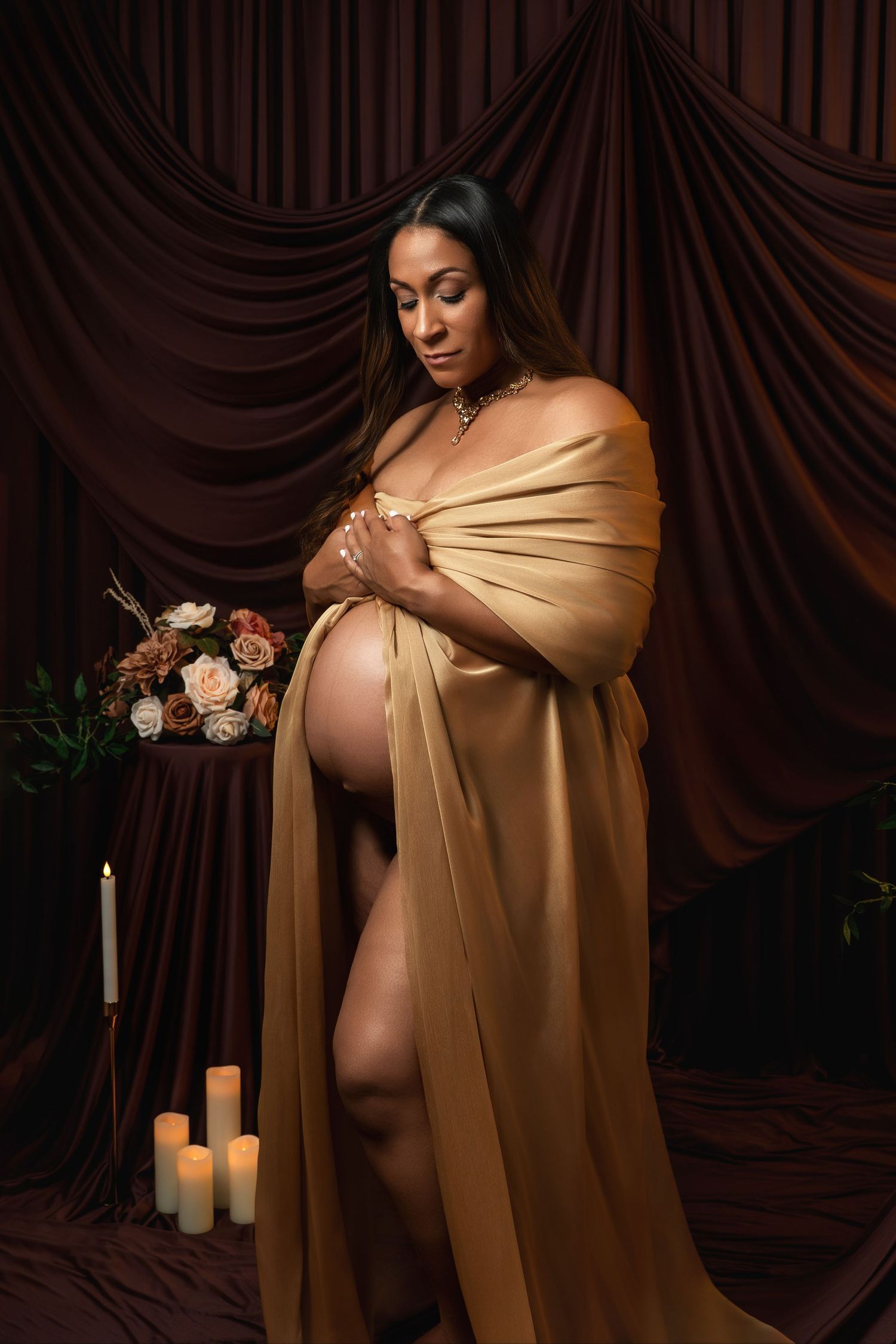 Artistic maternity portrait against dark brown backdrop with golden fabric draping and soft candlelight.