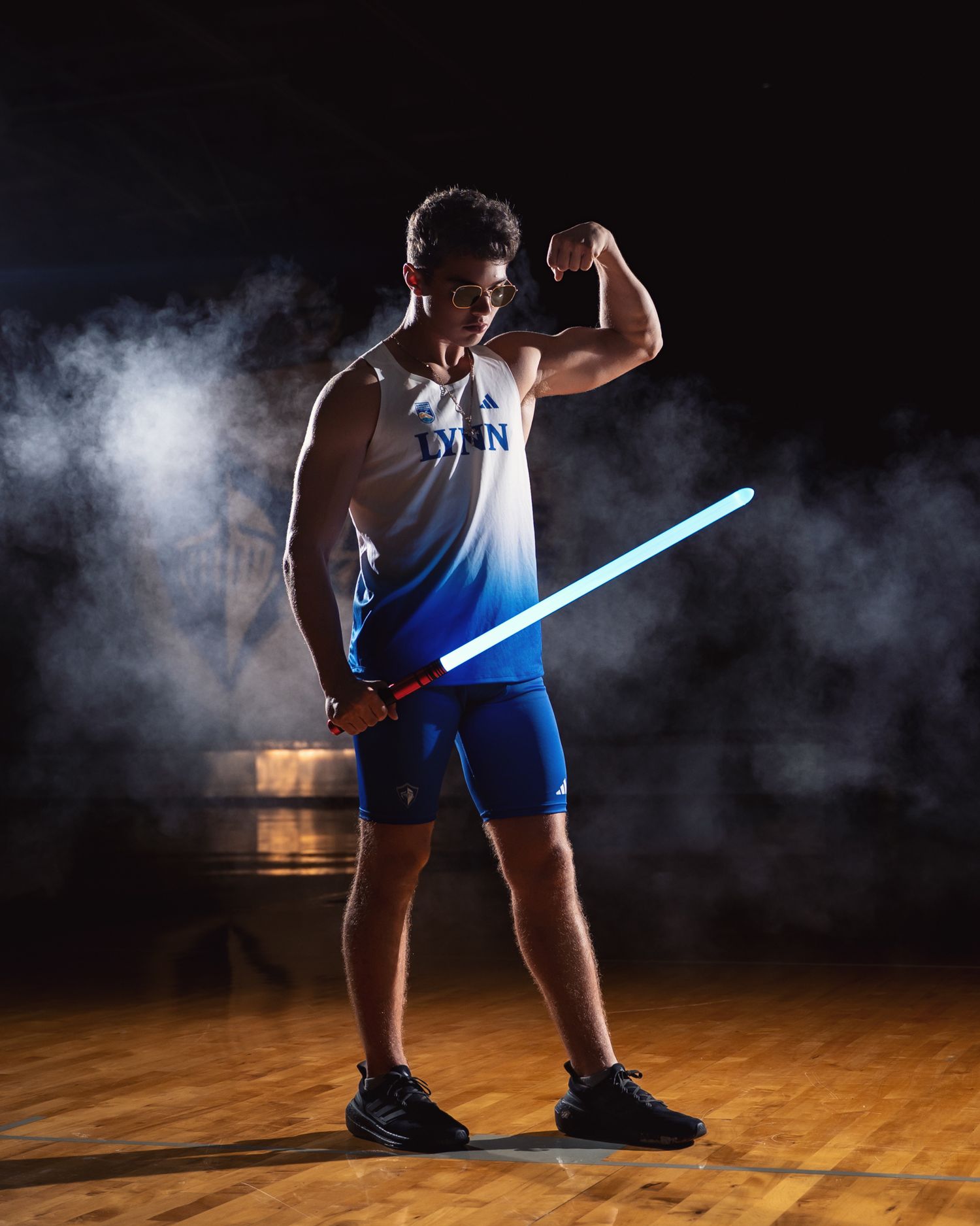 Lynn University athlete poses with glowing blue lightsaber in smoky gymnasium setting.