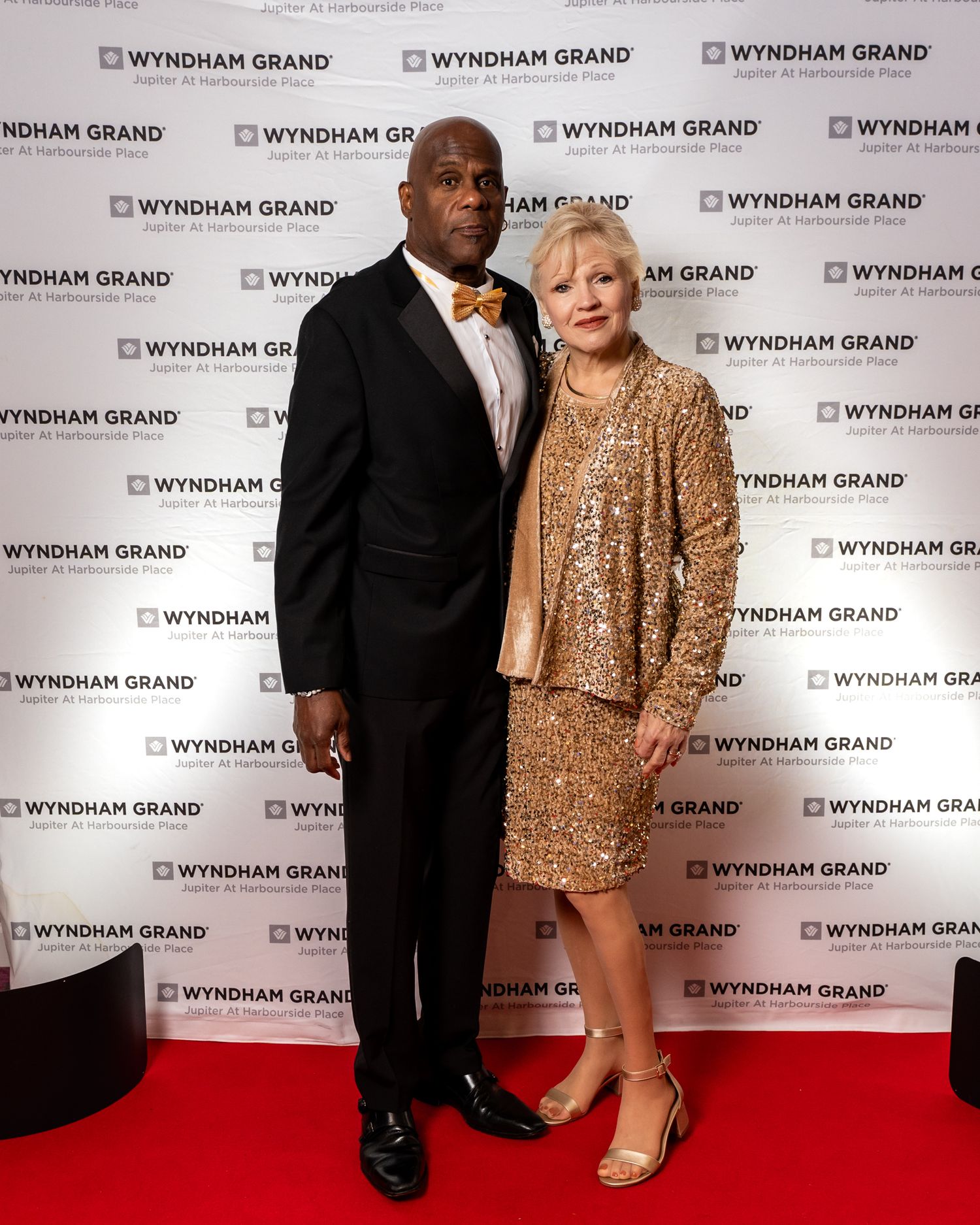 Two people posing at a Wyndham Grand step and repeat banner during a formal event.