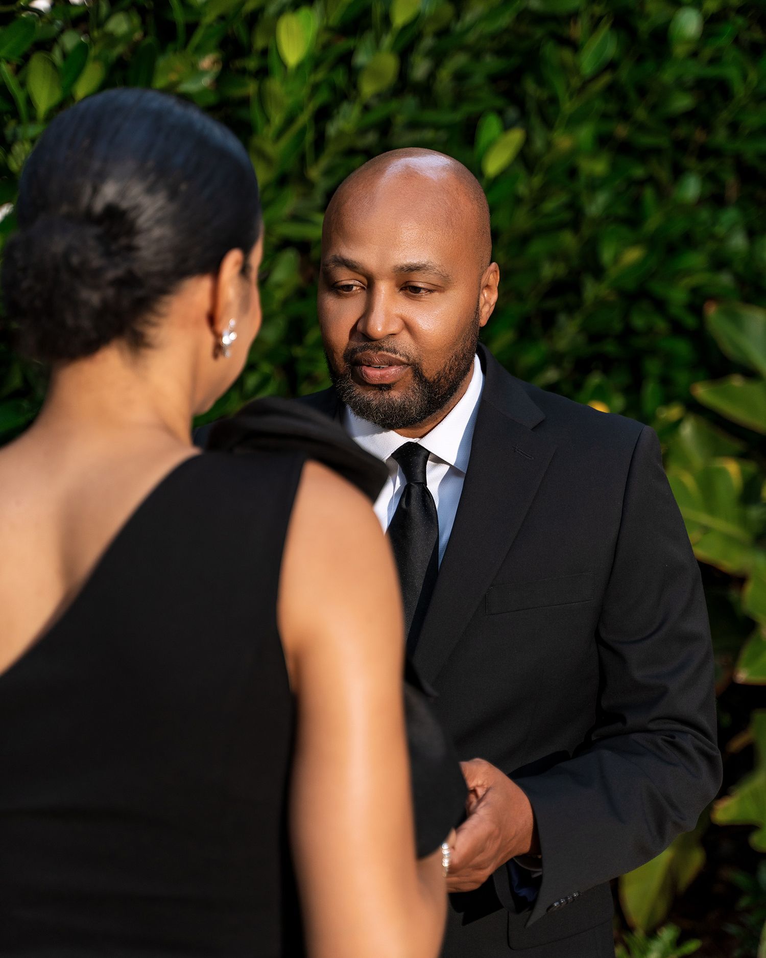 A couple shares a conversation in formal attire against a backdrop of green foliage.