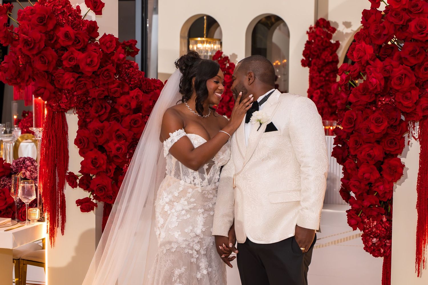 A couple shares an intimate moment at their wedding ceremony surrounded by vibrant red rose arrangements.