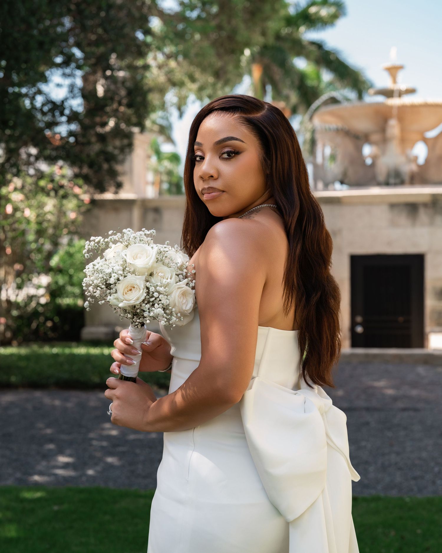 A bride in a white dress holding a bouquet of white flowers poses in an outdoor garden setting.