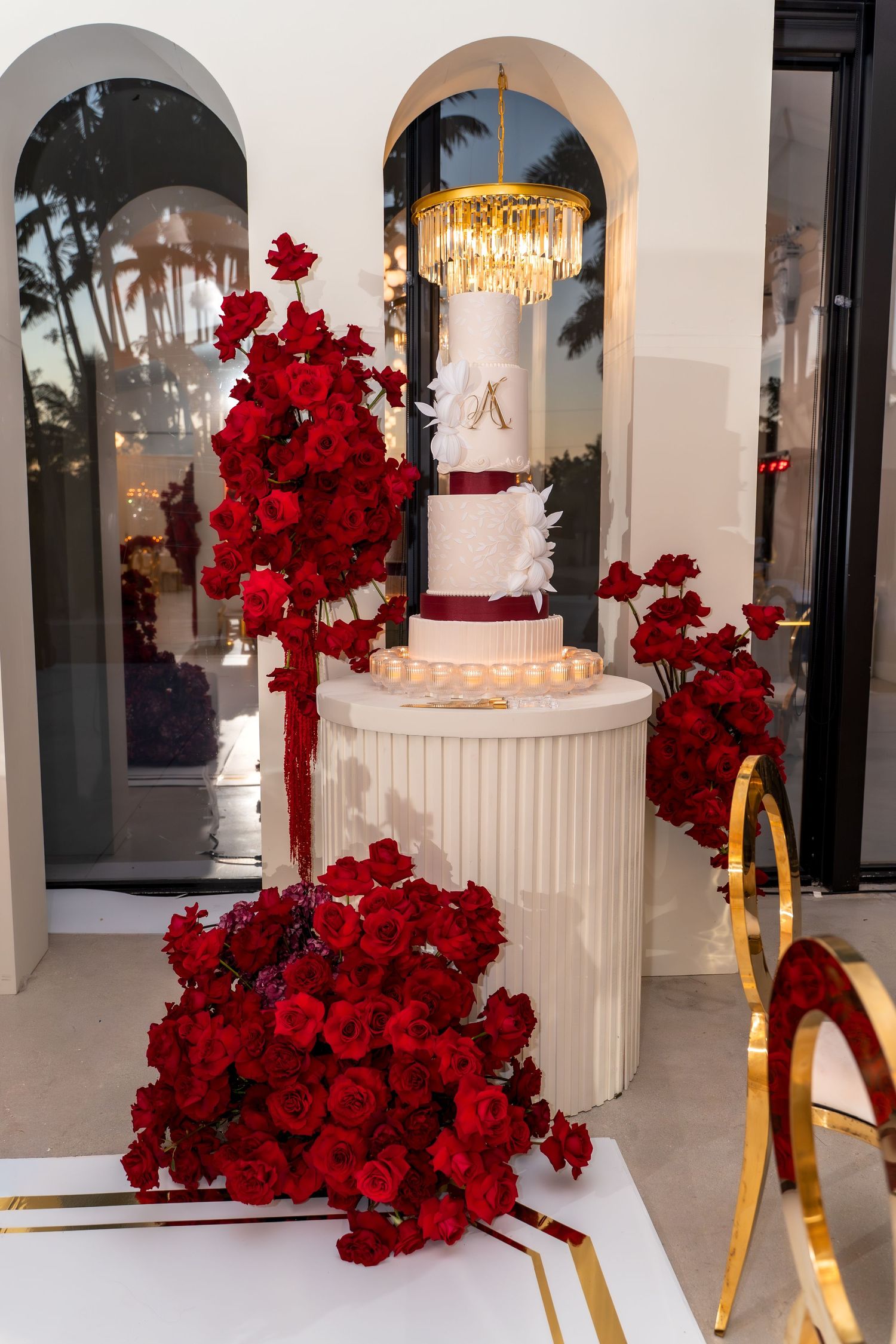 Elegant white wedding cake with red floral arrangements displayed on a pedestal under an arched doorway with chandelier.