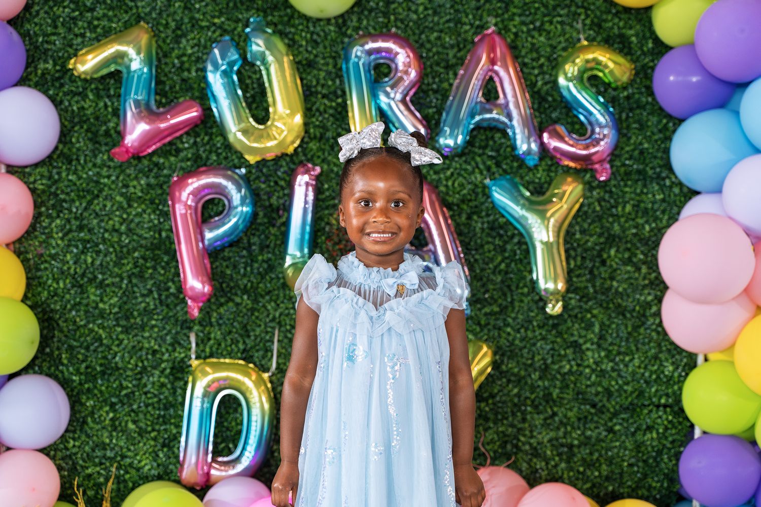 Colorful balloon display with 'LURA'S PLAY' text creates a festive backdrop against artificial grass.